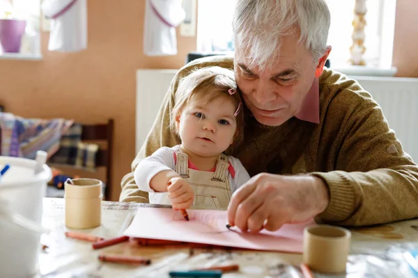 Menina pequena bonito bebê criança e bonito avô sênior pintura com lápis coloridos em casa. Neto e homem se divertindo juntos — Fotografia de Stock