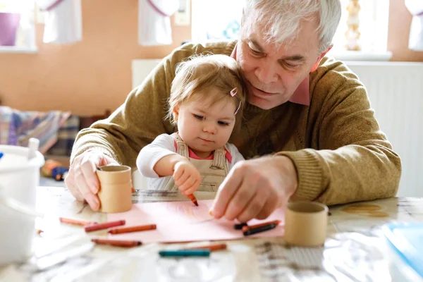 Schattig klein baby peuter meisje en knappe senior grootvader schilderij met kleurrijke potloden thuis. Kleinkind en man hebben plezier samen — Stockfoto