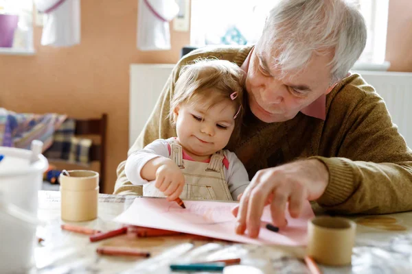 Χαριτωμένο μικρό κοριτσάκι και όμορφος ανώτερος παππούς ζωγραφική με πολύχρωμα μολύβια στο σπίτι. Εγγονός και άντρας διασκεδάζουν μαζί. — Φωτογραφία Αρχείου