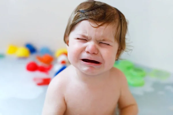 Söpö surullinen vauva tyttö ottaa vaahtoava kylpy kylpyammeessa. Itkevä lapsi leikkii kylpy kumi leluja — kuvapankkivalokuva