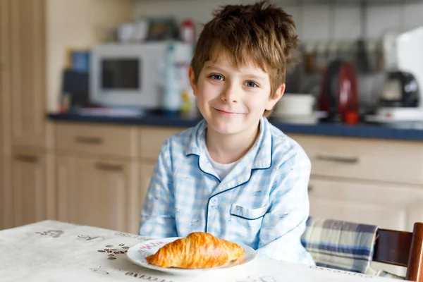 Счастливый маленький блондин, который ест свежий круассан на завтрак или обед. Здоровое питание для детей — стоковое фото