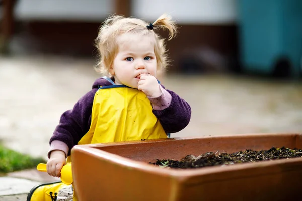 Cute adorable toddler girl playing with sand and shovel on spring day. Baby child wearing yellow boots and mud rain puddle pants. — Stock Photo, Image