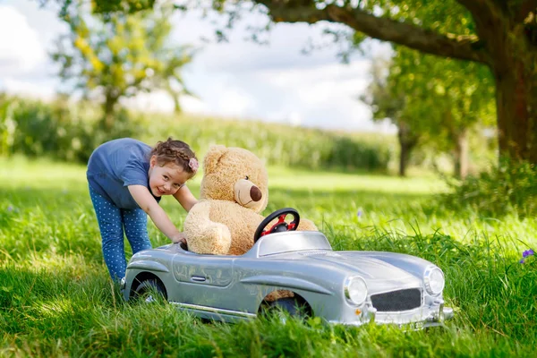 Pequena menina criança pré-escolar dirigindo carro de brinquedo grande e se divertindo com brincar com grande bea brinquedo de pelúcia — Fotografia de Stock