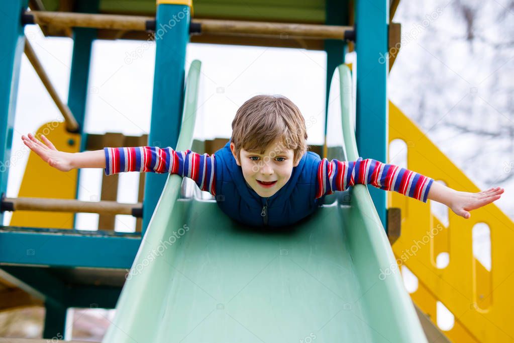 Happy blond kid boy having fun and sliding on outdoor playground