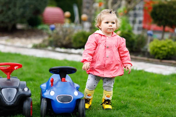 Niedliches kleines Mädchen, das mit zwei Spielzeugautos im Garten spielt. entzückendes Kleinkind, das Spaß hat. Mädchen in bunten Modekleidern. — Stockfoto