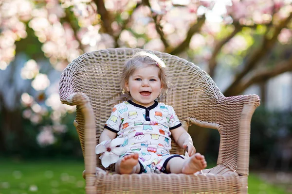 Söt liten baby flicka sitter på stora stol i trädgården. Vackra glada leende barn med blommande rosa magnoliaträd på bakgrund. — Stockfoto
