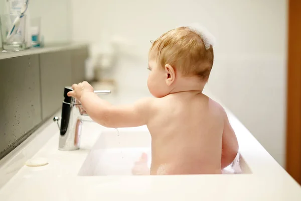 Милый очаровательный ребенок принимает ванну в раковине и играет с водой и пеной — стоковое фото