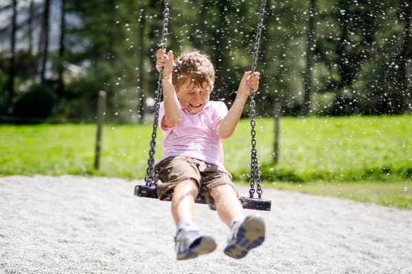 Funny kid boy having fun with chain swing on outdoor playground while being wet splashed with water — Stock Photo, Image