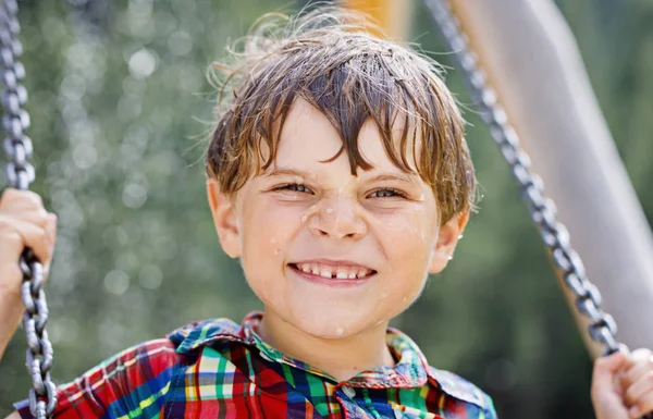 Lustiger kleiner Junge hat Spaß mit Kettenschaukel auf Spielplatz im Freien, während er nass mit Wasser bespritzt wird — Stockfoto