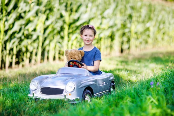 Pequena menina criança pré-escolar dirigindo carro de brinquedo grande e se divertindo com brincar com grande bea brinquedo de pelúcia — Fotografia de Stock