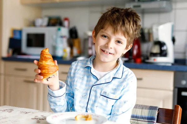 Happy little blond kid boy eating fresh croissant for breakfast or lunch. Healthy eating for children