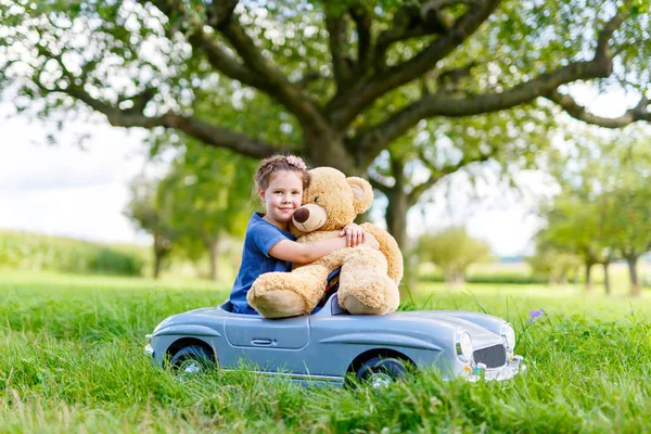 Pequeña niña preescolar conduciendo un gran coche de juguete y divirtiéndose jugando con un gran oso de peluche — Foto de Stock