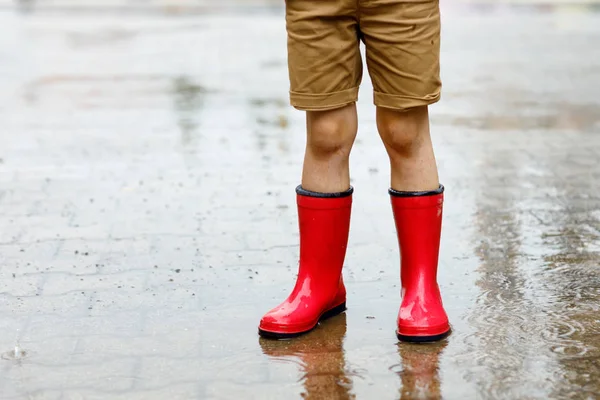 Niño con botas de lluvia rojas saltando en un charco . — Foto de Stock