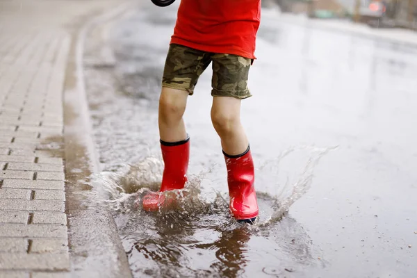 Niño con botas de lluvia rojas saltando en un charco . — Foto de Stock