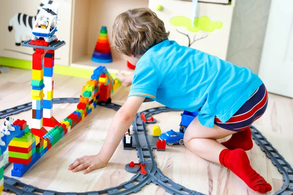 Menino loiro adorável brincando com blocos de plástico coloridos e criando estação de trem. Criança se divertindo com a construção de brinquedos ferroviários em casa — Fotografia de Stock