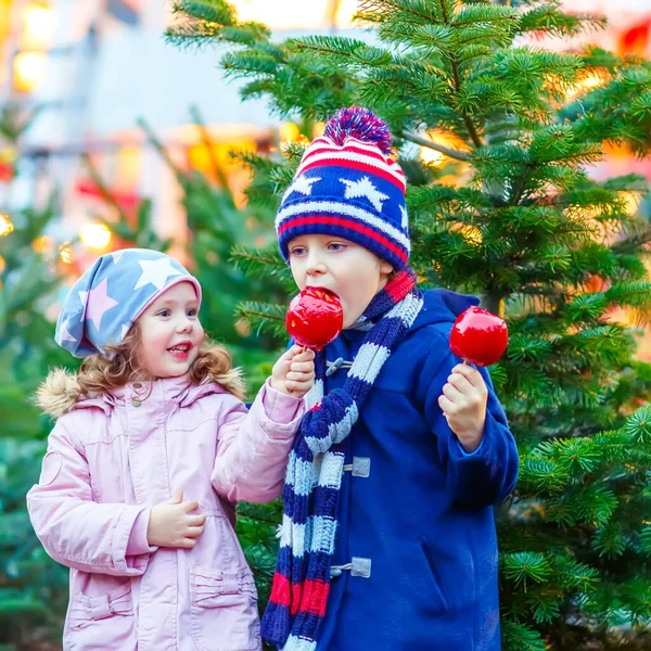 Dvě malé usmívající se děti, chlapec a dívka jíst slazené apple na vánoční trh. Šťastné děti v zimě módní oblečení s osvětlením na pozadí. Srandovní sestra a bratr. — Stock fotografie