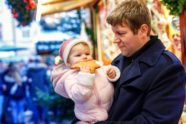 Midden leeftijd vader baby dochter in de buurt van zoete stand met peperkoek en noten te houden. Gelukkige familie op de kerstmarkt in Duitsland. Schattig meisje eten cookie met de naam "Lebkuchen". Viering kerstvakantie. — Stockfoto