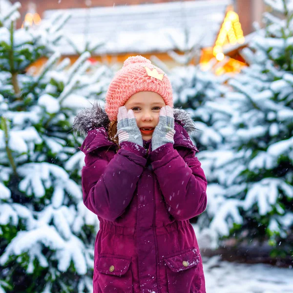 Söta lilla leende kid flicka på julgran marknaden. Glada barn i vinterkläder att välja xmas tree på xmas marknaden med ljus på bakgrunden på vintern snö dag. — Stockfoto