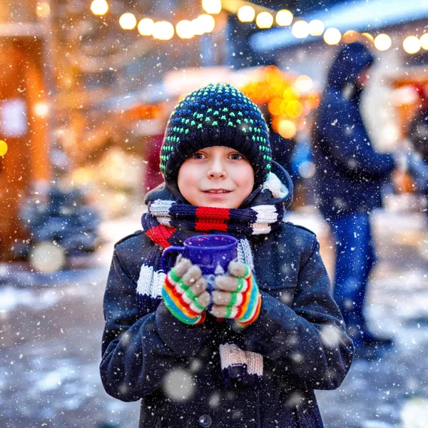 Kleine schattige jongen drinken hete kinderen punch of chocolade op de Duitse kerstmarkt. Gelukkig kind op traditionele familiemarkt in Duitsland, lachende jongen in kleurrijke winterkleding — Stockfoto