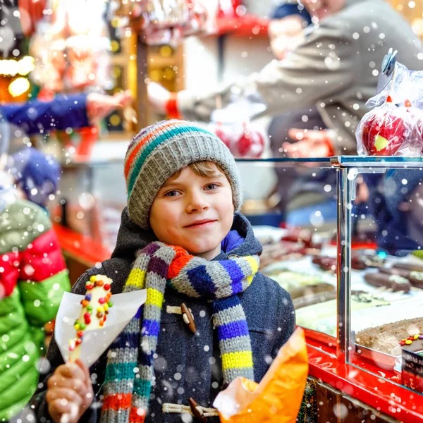 Niño pequeño, niño lindo comiendo plátanos cubiertos de chocolate, marshmellows y coloridos espolvoreos cerca de puesto dulce con pan de jengibre y nueces. Niño feliz en el mercado de Navidad en Alemania . — Foto de Stock