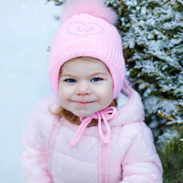 Retrato de una niñita caminando al aire libre en invierno. Lindo niño comiendo dulces piruletas dulces. Niño divirtiéndose en el frío día de nieve. El uso de ropa de bebé caliente de color rosa y sombrero con bobbles. — Foto de Stock