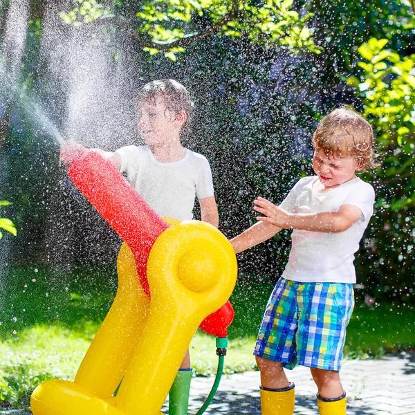 Two little kids boys playing with a garden hose sprinkler on hot and sunny summer day. Children having fun with sprinkling water, drops rain. Outdoors leisure wth water for friends. Rain rubber boots. — Stock Photo, Image