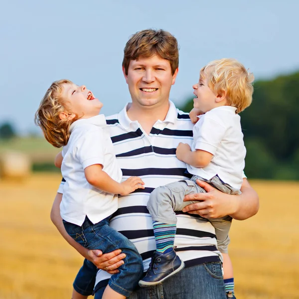 Happy family enjoying sunset in wheat field. Beautiful young man with two adorable toddler kids boys. Father holding two children, twins on arms on meadow on a sunny evening. Dad and sons. Outdoors — Stock Photo, Image