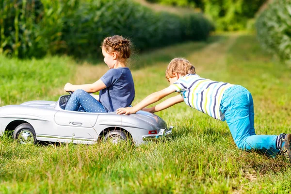 Dos niños felices jugando con un gran coche de juguete viejo en el jardín de verano, al aire libre. Un chico conduciendo un auto con una niña dentro. Riendo y sonriendo niños. Familia, infancia, concepto de estilo de vida —  Fotos de Stock
