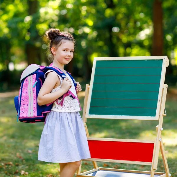 Glad liten unge flicka står vid skrivbord med ryggsäck eller axelväska. Schoolkid första dagen av elementär klass. Tillbaka till skolan-konceptet. Friska bedårande barn utomhus, i grön park — Stockfoto