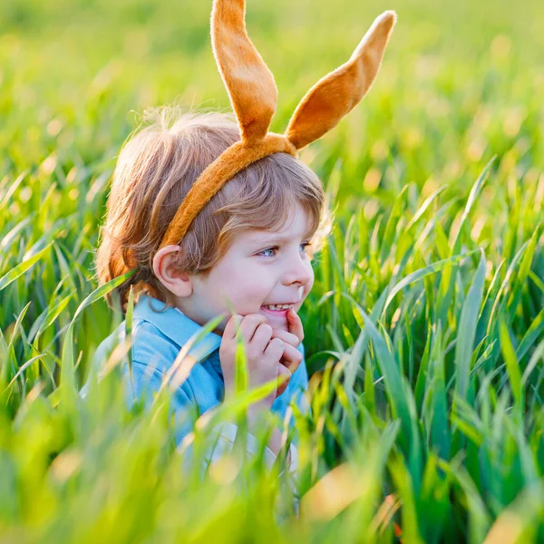 Niño lindo con orejas de conejo divirtiéndose con la tradicional caza de huevos de Pascua en el cálido día soleado, al aire libre. Celebrando las vacaciones de Pascua. Encontrar niños pequeños, huevos coloridos en hierba verde — Foto de Stock