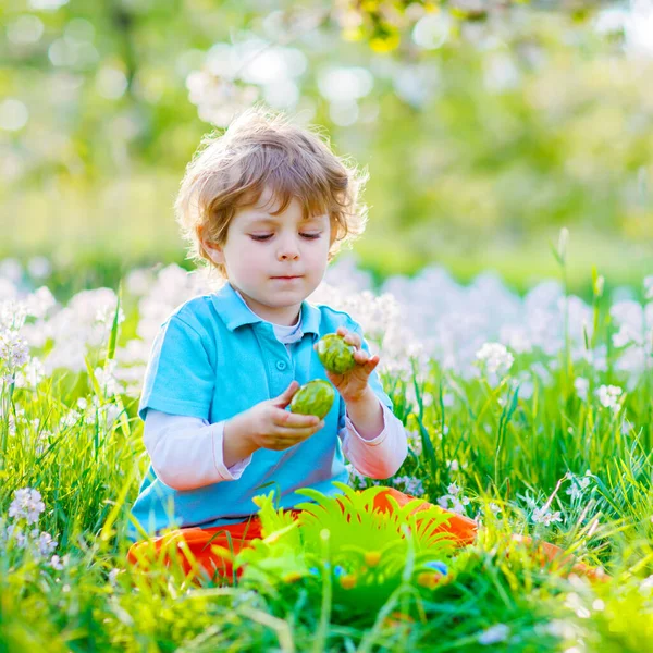 Ragazzino carino con orecchie da coniglio che si diverte con le tradizionali uova di Pasqua a caccia nelle calde giornate di sole, all'aperto. Festeggiamo la festa di Pasqua. Trovare bambino, uova colorate in erba verde — Foto Stock