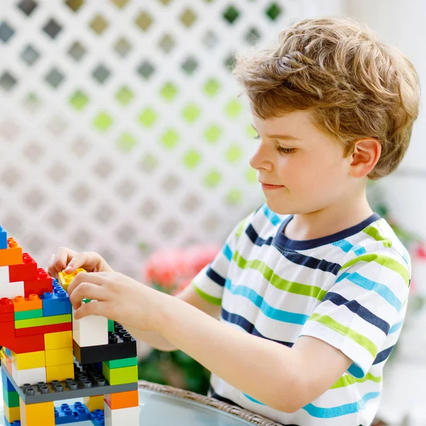 Маленький блондин играет с кучей разноцветных пластиковых блоков. Очаровательный дошкольный мальчик в разноцветной рубашке и с удовольствием строит большой замок и создает дом — стоковое фото