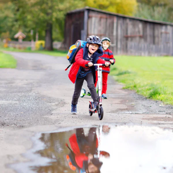 Mignon petit garçon de l'école chevauchant sur scooter push sur le chemin de ou vers l'école. Écolier de 7 ans conduisant dans une flaque d'eau de pluie. drôle enfant heureux dans des vêtements de mode colorés et avec casque. — Photo