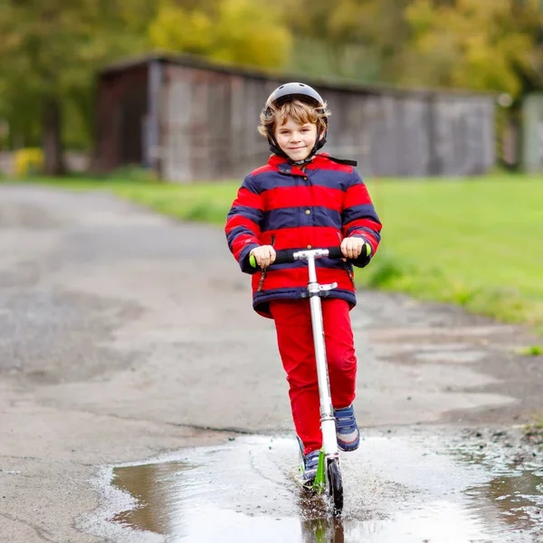 Cute little school kid boy riding on push scooter on the way to or from school. Schoolboy of 7 years driving through rain puddle. funny happy child in colorful fashion clothes and with helmet. — Stock Photo, Image