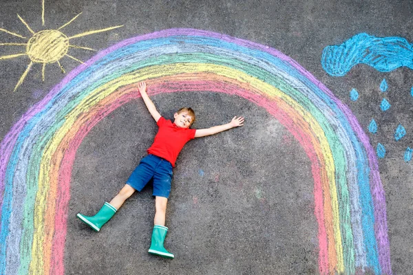 Niño feliz en botas de goma con sol de arco iris y nubes con gotas de lluvia pintadas con tiza de colores en el suelo o asfalto en verano. Ocio creativo para niños al aire libre en verano — Foto de Stock