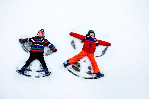 Dos hermanos pequeños niños con ropa de invierno de colores haciendo ángel de nieve, acostado en la nieve. Activo ocio al aire libre con niños en invierno. Hermanos felices con sombrero caliente, guantes, moda de invierno. —  Fotos de Stock