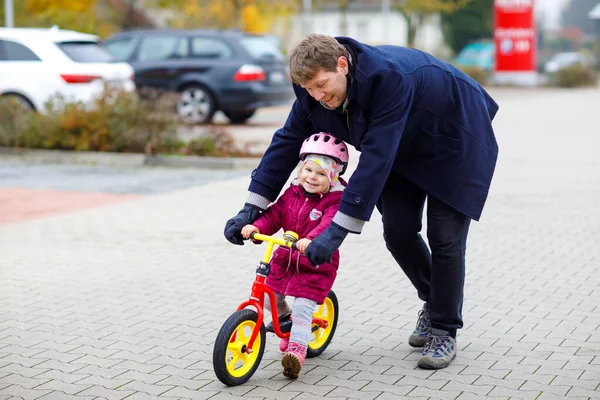 ランバランスバイクに乗って安全ヘルメットに乗ってかわいい幼児の女の子。中年の父親は幸せな健康的な素敵な赤ちゃんの娘に自転車を学ぶことを楽しんで教えています。屋外で活動する家族. — ストック写真