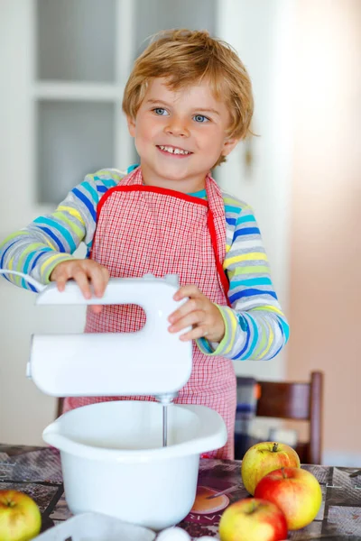 Aranyos kis szőke óvodás fiú almatortát és muffint süt a háztartási konyhában. Vicces szép egészséges gyermek szórakozás dolgozik mixer, liszt, tojás, gyümölcsök. Kis segítség a házban. — Stock Fotó