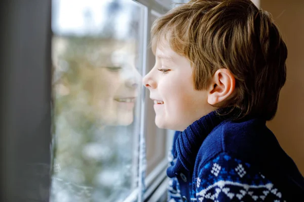 Glad bedårande liten pojke sitter nära fönstret och tittar ut på snö på juldagen eller morgonen. Leende friska barn fascinerad observera snöfall och stora snöflingor — Stockfoto