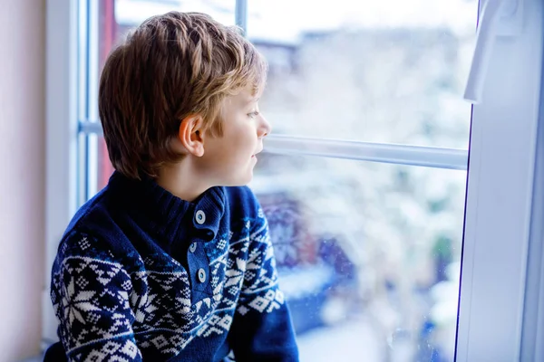 Szczęśliwy uroczy chłopiec siedzi w pobliżu okna i patrzy na zewnątrz na śnieg w Boże Narodzenie lub rano. Uśmiechnięte zdrowe dziecko fascynowane obserwacją opadów śniegu i dużych płatków śniegu — Zdjęcie stockowe