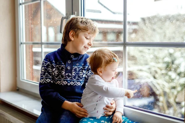 Happy adorable kid boy and cute baby girl sitting near window and looking outside on snow on Christmas day or morning. Smiling children, siblings, little sister and brother looking on winter snowfall. — Stock Photo, Image