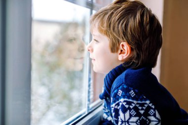 Mutlu sevimli çocuk pencerenin yanında oturuyor ve Noel sabahı ya da sabahleyin dışarıda karları seyrediyor. Gülümseyen sağlıklı çocuk, karları ve büyük kar tanelerini izlerken büyülenir.