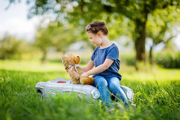 Kleine Vorschulmädchen fahren große alte Spielzeugautos und haben Spaß beim Spielen mit großen Plüschbären, im Freien. Kind genießt warmen Sommertag in der Naturlandschaft. Mädchen fährt Auto — Stockfoto