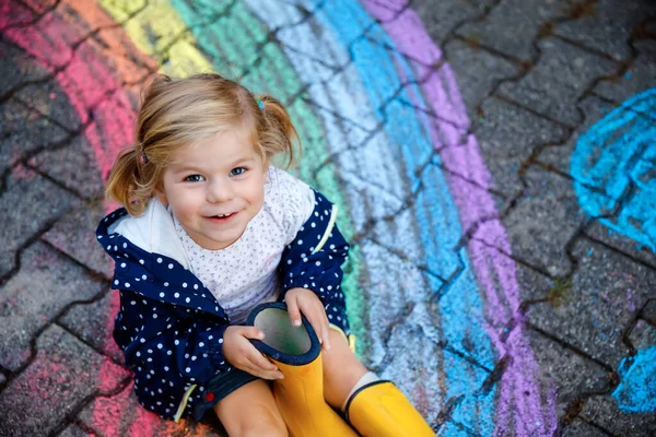 Menina pequena feliz em botas de borracha com sol arco-íris e nuvens com chuva pintada com giz colorido no chão ou asfalto no verão. Criança bonita se divertindo. lazer criativo — Fotografia de Stock