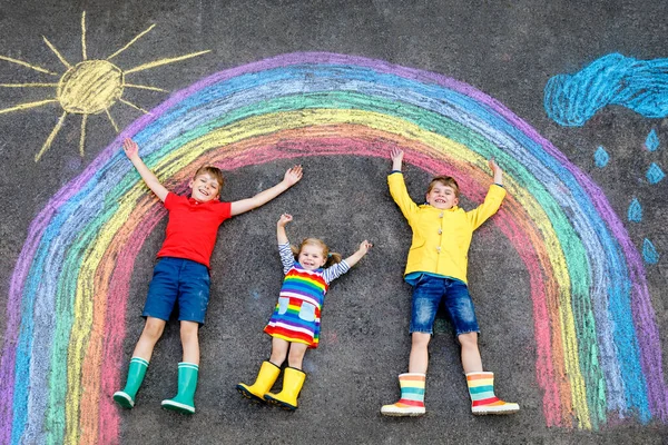 Drei kleine Kinder, zwei Schulkinder Jungen und ein Kleinkind amüsieren sich mit Regenbogenbildern mit bunten Kreiden auf Asphalt. Geschwister in Gummistiefeln malen auf dem Boden und spielen zusammen. — Stockfoto