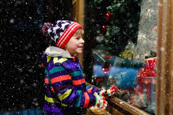 Roztomilý malý, zdravý školák na vánočním trhu. Vtipné šťastné dítě v módě zimní oblečení, aby okno nakupování zdobené dárky, vánoční strom. Padá sníh, padá sníh — Stock fotografie