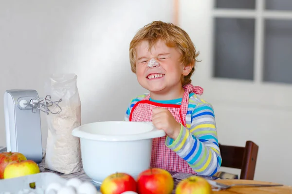 Bonito pequeno menino pré-escolar loiro feliz cozinhando bolo de maçã e muffins na cozinha doméstica. Engraçado adorável criança saudável se divertindo com o trabalho com misturador, farinha, ovos, frutas. Pequeno ajudante dentro de casa — Fotografia de Stock