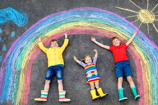 Drei kleine Kinder, zwei Schulkinder Jungen und ein Kleinkind amüsieren sich mit Regenbogenbildern mit bunten Kreiden auf Asphalt. Geschwister in Gummistiefeln malen auf dem Boden und spielen zusammen. — Stockfoto