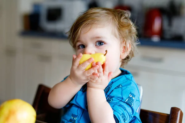 かわいい愛らしい幼児の女の子は新鮮な梨を食べています。1年間の果物を保持の空腹幸せな赤ちゃん。家庭用キッチンの女の子、健康的な食事の軽食を持っている。笑顔ブロンドの子供 — ストック写真