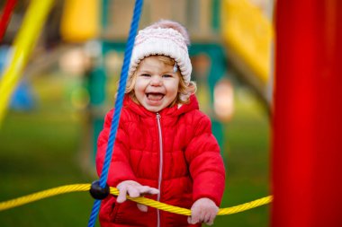 Küçük tatlı kız oyun alanında eğleniyor. Mutlu küçük çocuk tırmanışı, farklı ekipmanların üzerinde sallanmak ve kaymak. Renkli giysiler içinde soğuk bir günde. Çocuklar için aktif açık hava oyunu